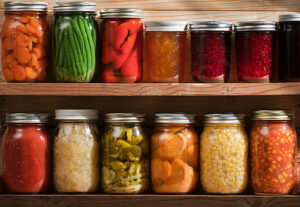 Subject: Two wooden shelves holding a variety of canned vegetables and fruits, lined up in rows of glass jars. Food staples canned include jellies, sauces, or slices of carrots, green beans, tomatoes, corn, sweet potatoes, sauerkraut, roasted red peppers, dill pickles, raspberry jam, orange marmalade, grape jelly, and a tomato and corn soup.