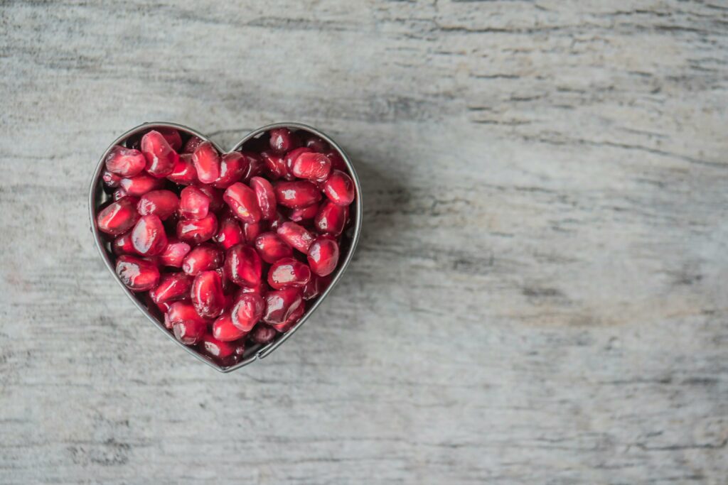 heart shaped bowl of pomegranate seeds against a gray toned wood background